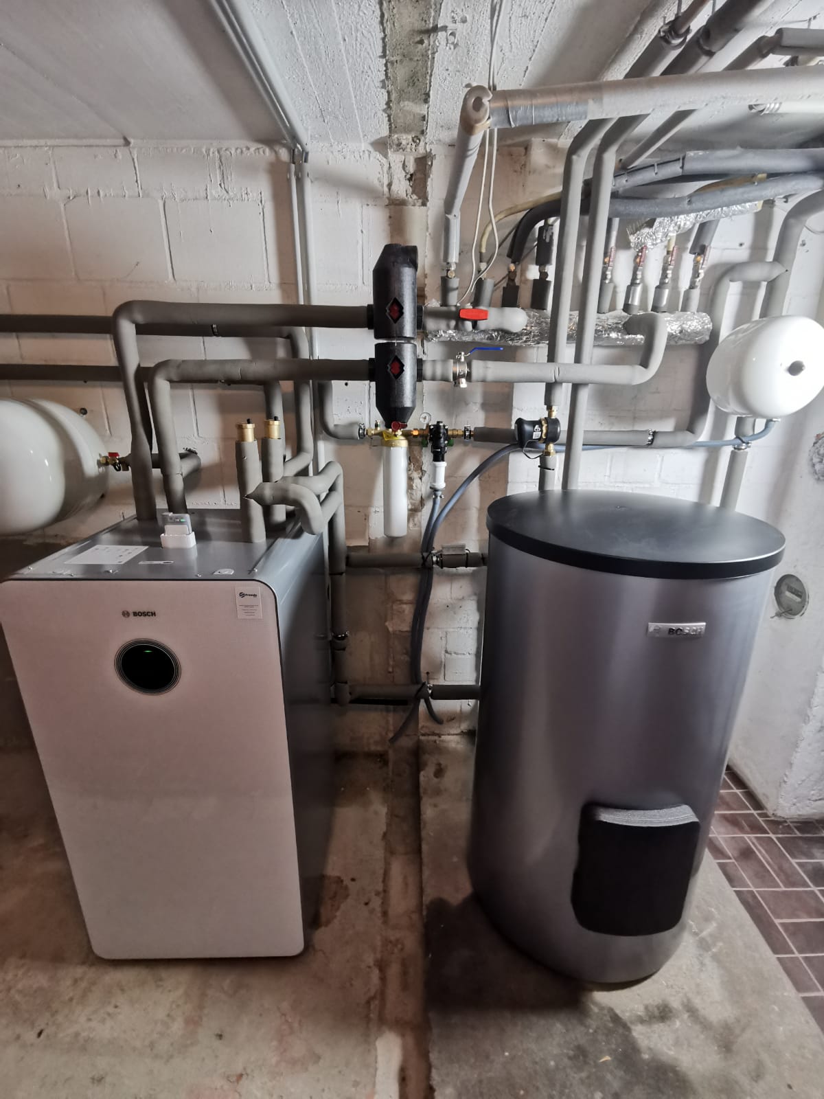 Wärmepumpen-Inneneinheit von Bosch mit Pufferspeicher, angeschlossen an ein komplexes Rohrsystem in einem sauberen Technikraum.